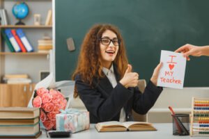 smiling-showing-thumbs-up-young-female-teacher-received-postcard-sitting-desk-with-school-tools-classroom